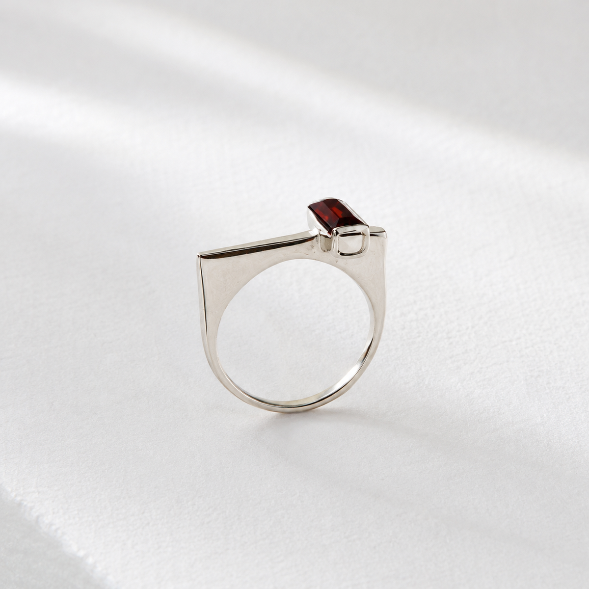 A silver ring with a red gemstone, possibly a garnet, is displayed against a white background.