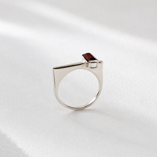 A silver ring with a red gemstone, possibly a garnet, is displayed against a white background.