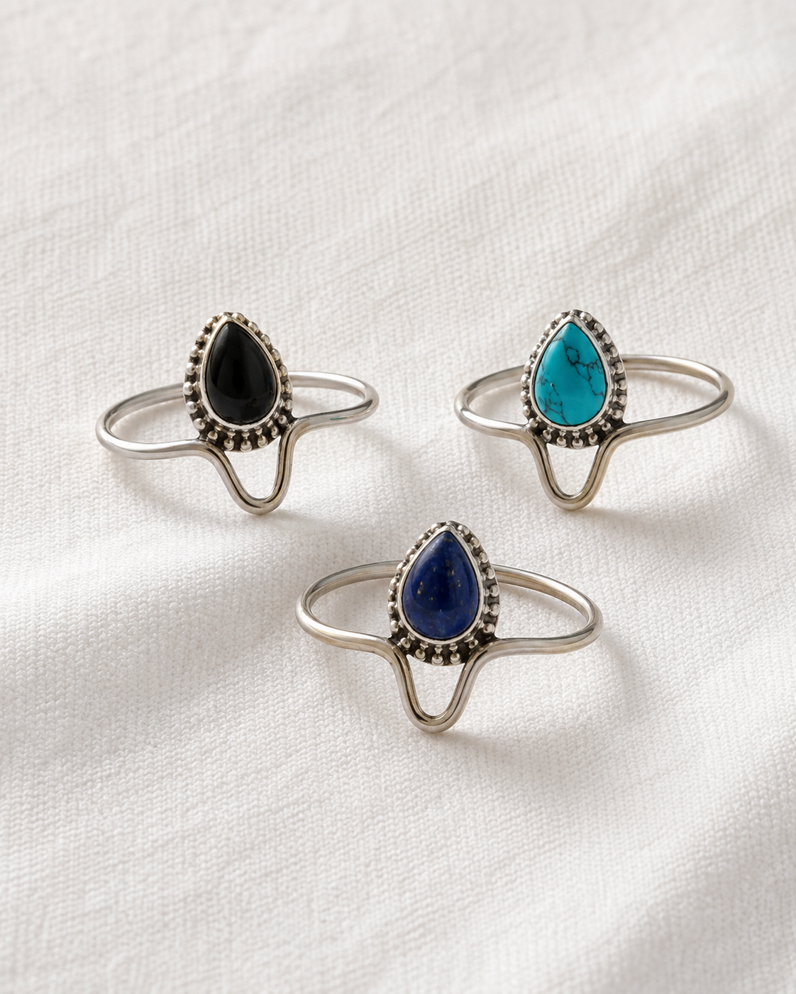 Three silver ring designs with gemstones, including a black stone, a blue stone, and a turquoise stone, are displayed on a white fabric background.