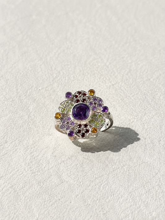 A silver ring with a large purple gemstone surrounded by smaller gemstones in various colors.