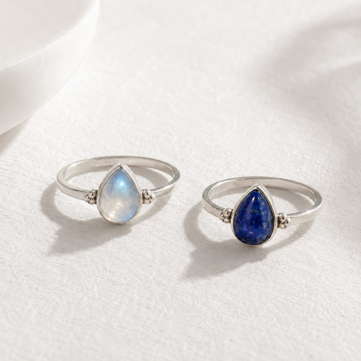 Two silver rings with gemstones, one with a blue gemstone and the other with a white gemstone, are displayed on a white surface.