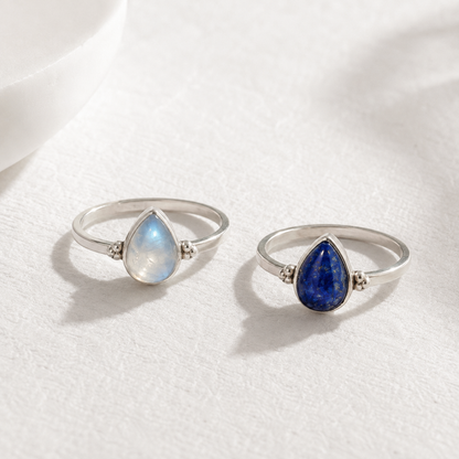 Two silver rings with gemstones, one with a blue gemstone and the other with a white gemstone, are displayed on a white surface.
