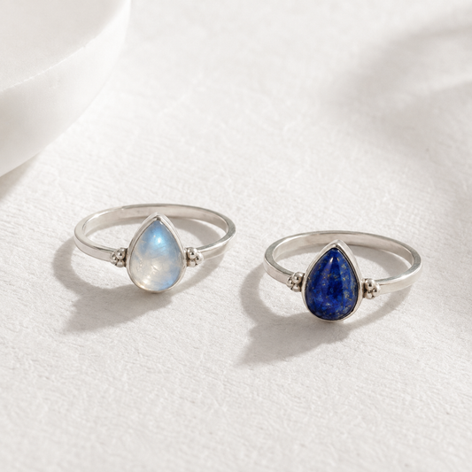 Two silver rings with gemstones, one with a blue gemstone and the other with a white gemstone, are displayed on a white surface.